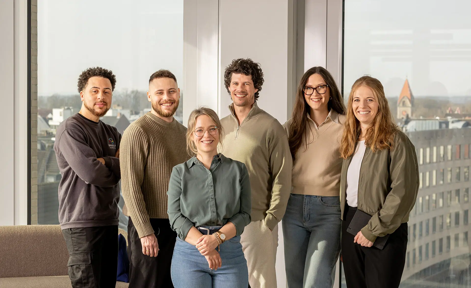 Teamfoto von 6 freundlich schauenden Personen in einem hellen Raum mit hohen Fenstern